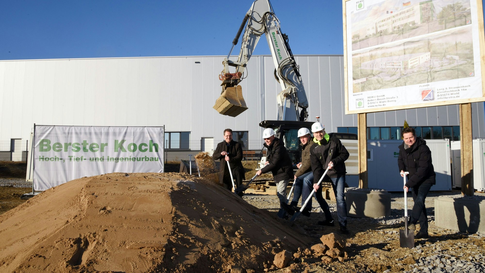 Fünf Männer hantieren mit Spaten vor einem Bagger.