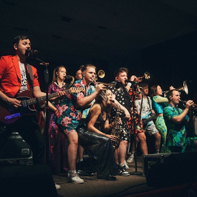 Auf der Bühne begeistert Druckluft das Publikum mit einer coolen Show und flotten Brassrhythmen. In der Mitte Interviewpartner Florian Hertel im Schwarzen Outfit mit weißen Sternen.