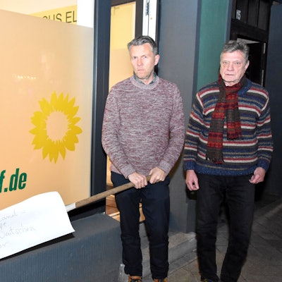 Auf dem Foto sind die Elsdorfer Grünen-Politiker Michael Broich und Hartmut Bergheim vor dem Büro ihrer Fraktion zu sehen. Broich hält die Harke in den Händen.