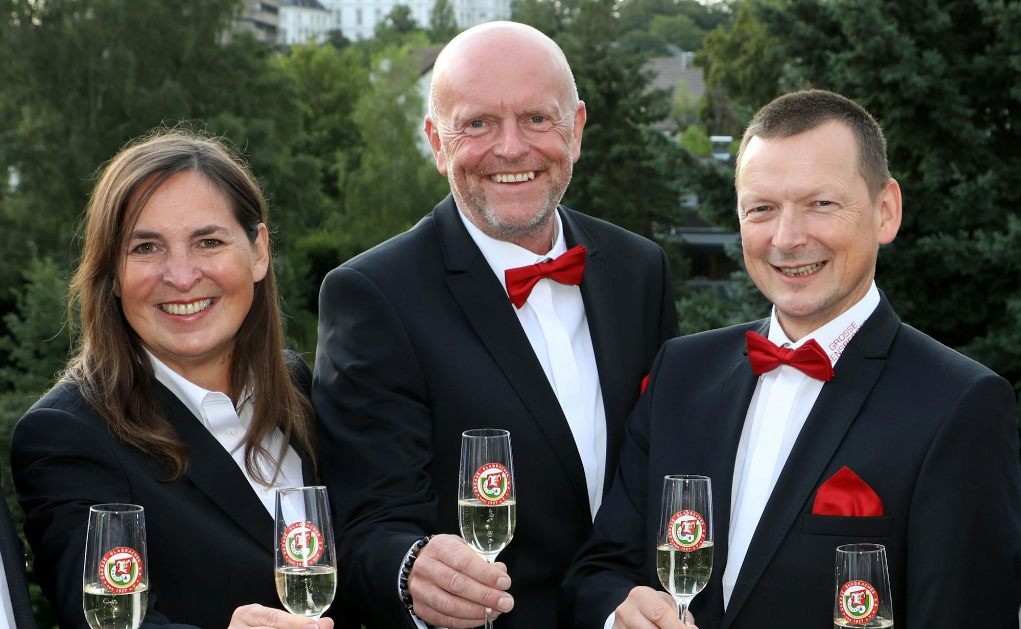 Drei Menschen stehen mit Sektgläsern vor einem grünen Garten.