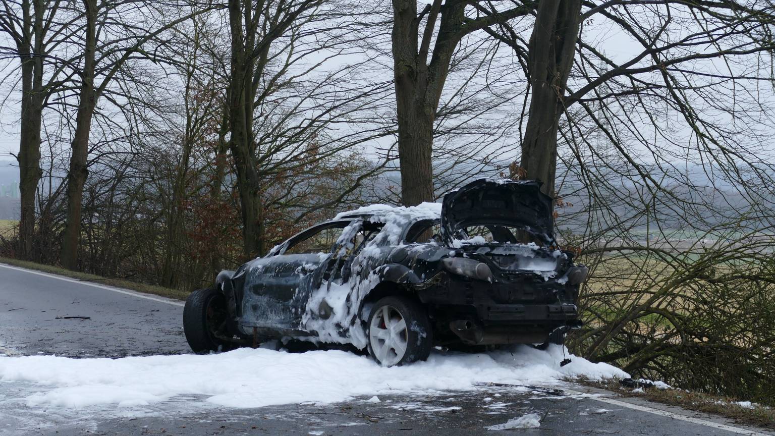 Ein ausgebranntes Auto steht mit Löschschaum bedeckt auf einer Straße.