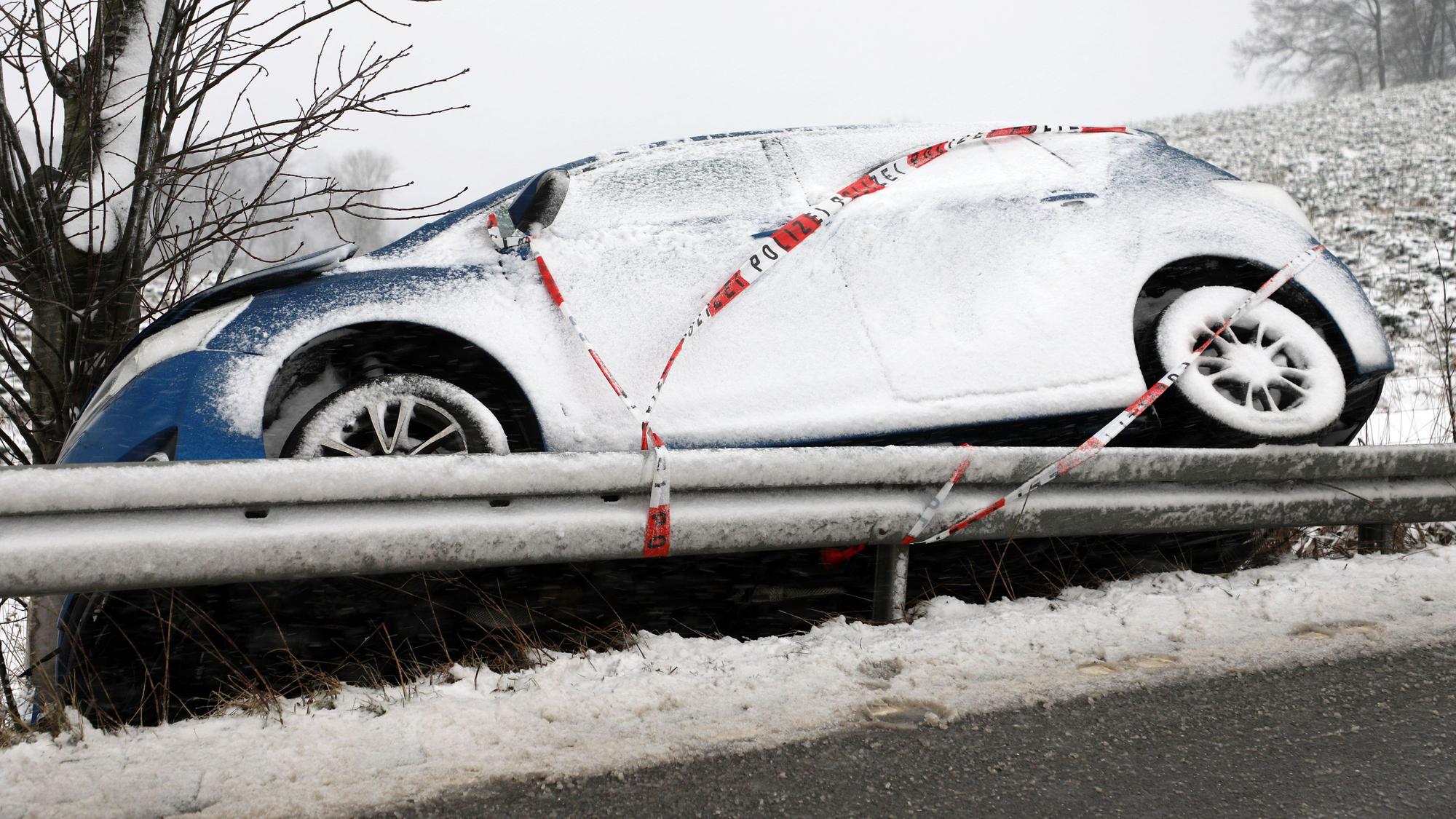 Ein Auto mit Polizei-Absperrband steht aufgebockt auf einer Leitplanke vor einem Baum. Schnee liegt auf dem Auto.
