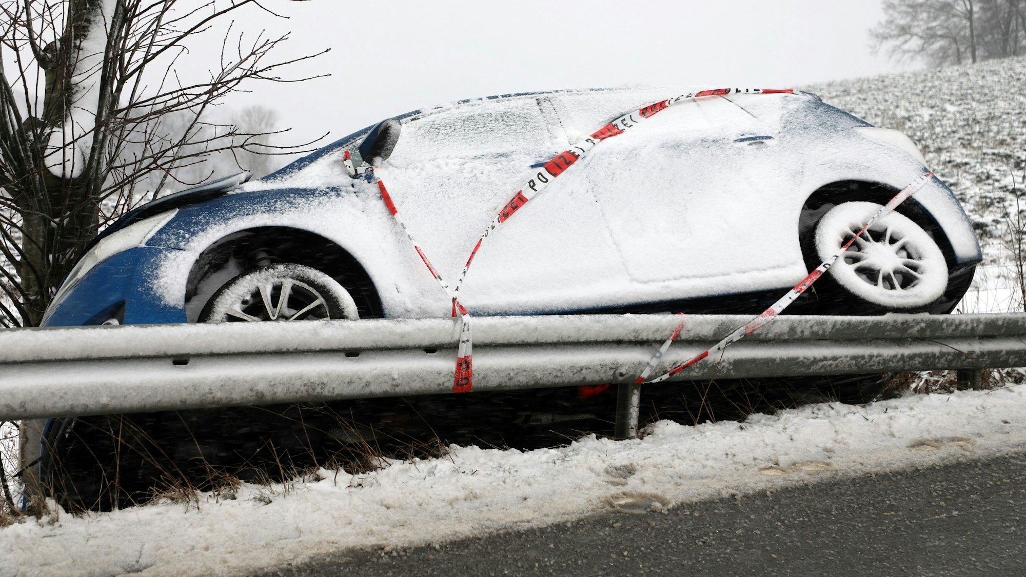 Ein Auto mit Polizei-Absperrband steht aufgebockt auf einer Leitplanke vor einem Baum. Schnee liegt auf dem Auto.