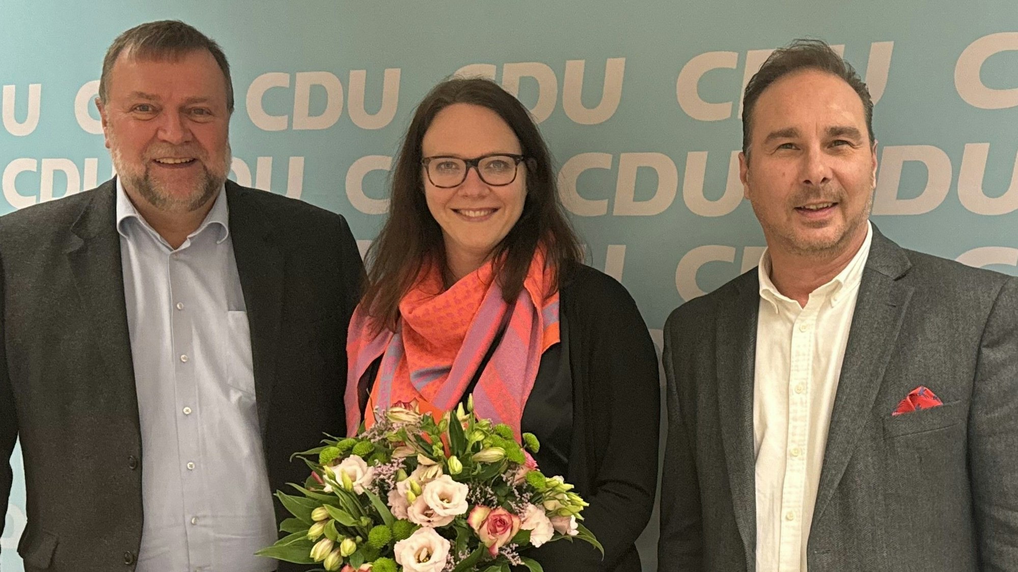 Zwei Männer im grauenSakko stehen links und rechts einer Frau mit Brille und rosa Halstuch. Die Frau hat einen Blumenstrauß in der Hand.