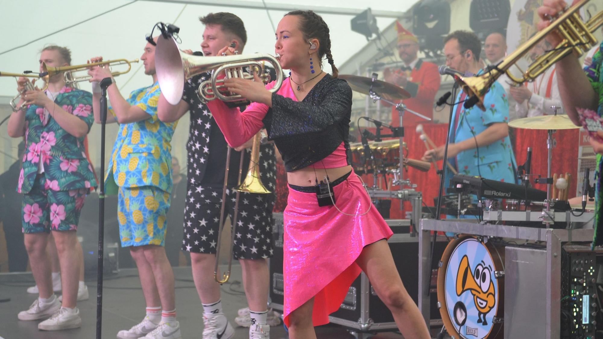Die Musikerinnen und Musik der Band Druckluft stehen in ihrer bunten Kleidung auf der Bühne, unter anderem mit Posaunen und Trompeten.