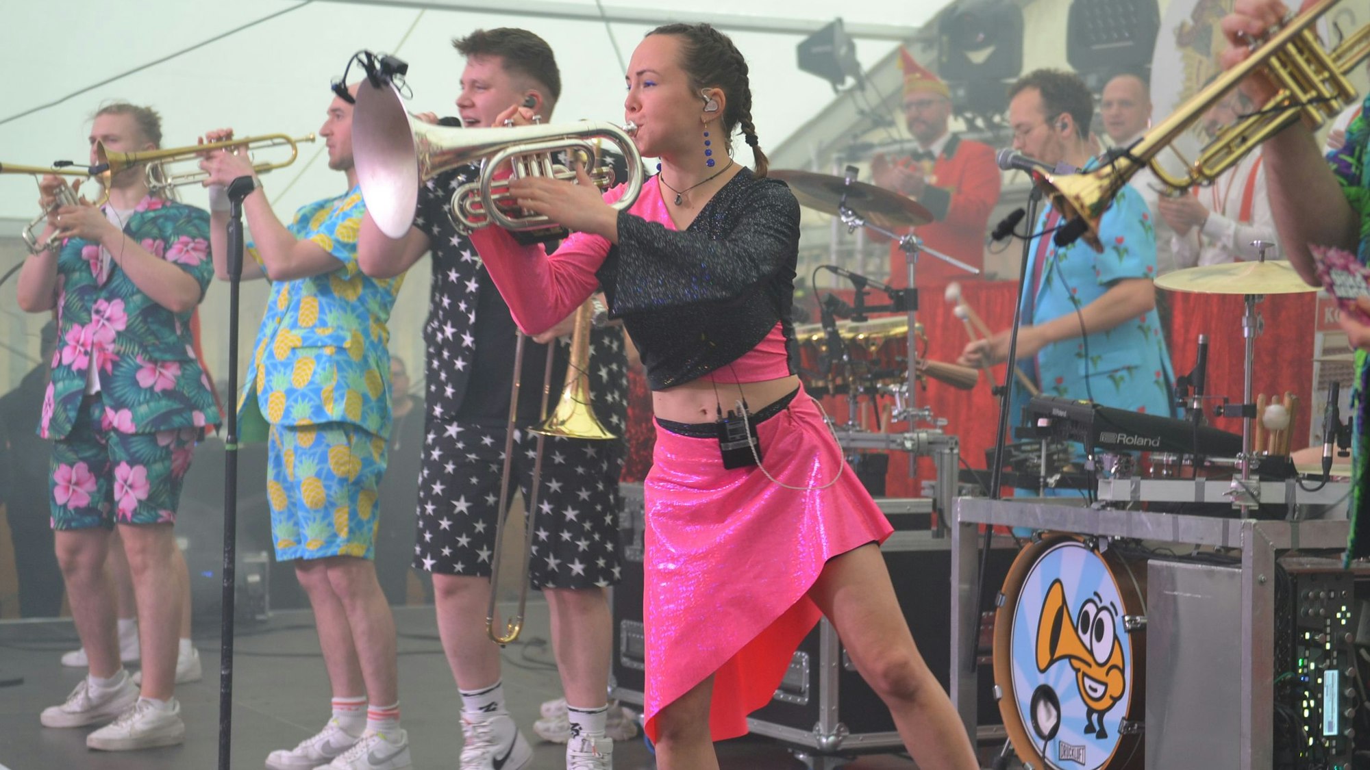 Die Musikerinnen und Musik der Band Druckluft stehen in ihrer bunten Kleidung auf der Bühne, unter anderem mit Posaunen und Trompeten.