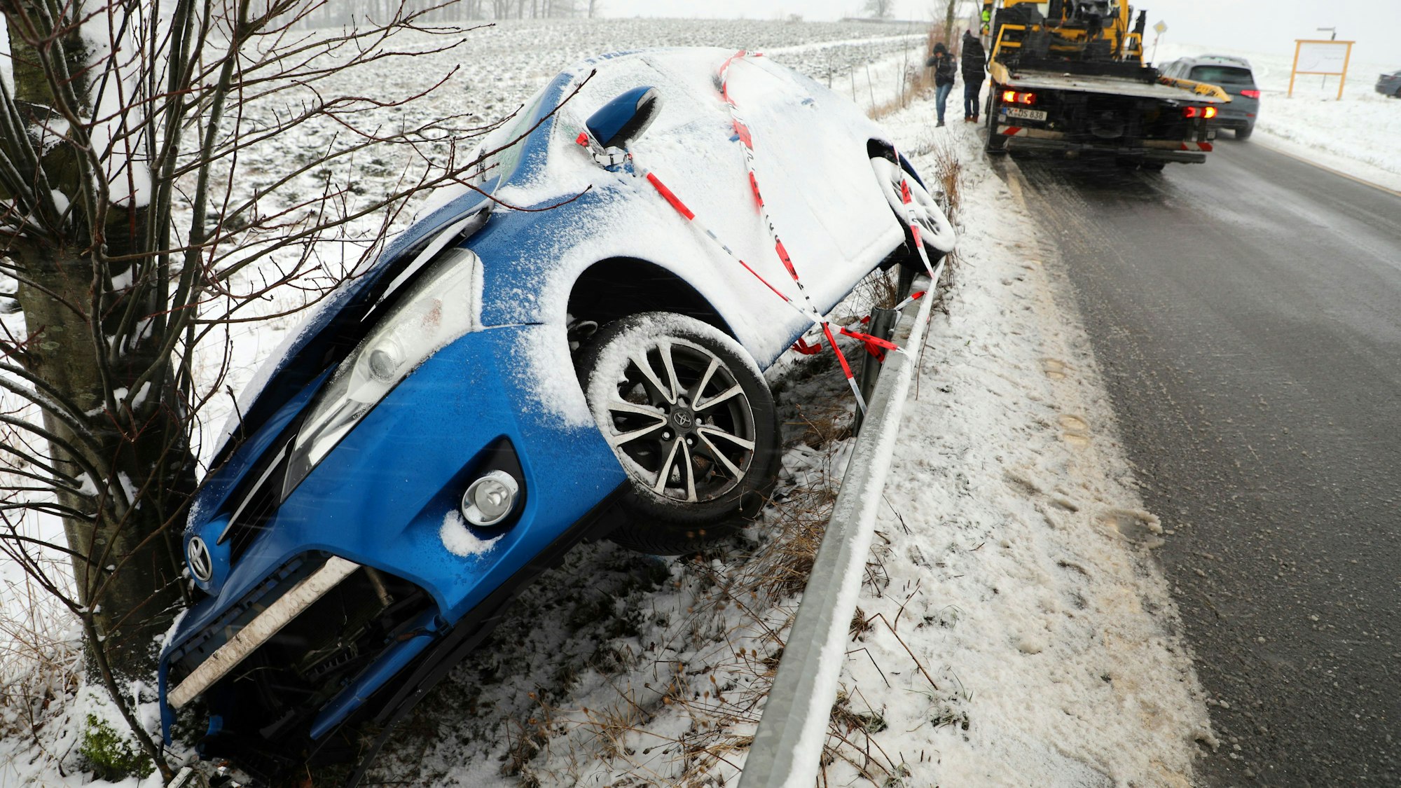 Ein zugeschneites Auto steht beschädigt auf einer Leitplanke vor einem Baum neben einer Straße.