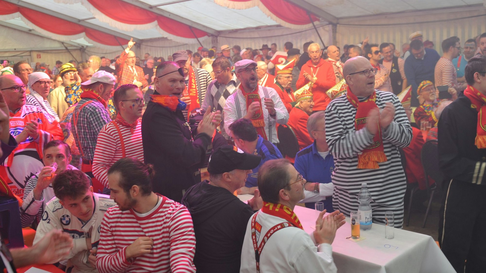 Kostümierte Männer sitzen und stehen dicht an dicht im Karnevalsfestzelt, viele klatschen und singen.