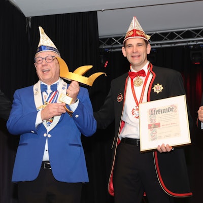 Drei Karnevalisten stehen beisammen. Der Mann in der Mitte hält eine Auszeichnung in Form einer goldenen Narrenkappe in den Händen.