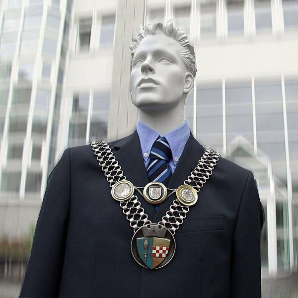 Eine Puppe trägt die Amtskette des Bürgermeisters vor einem Rathaus.