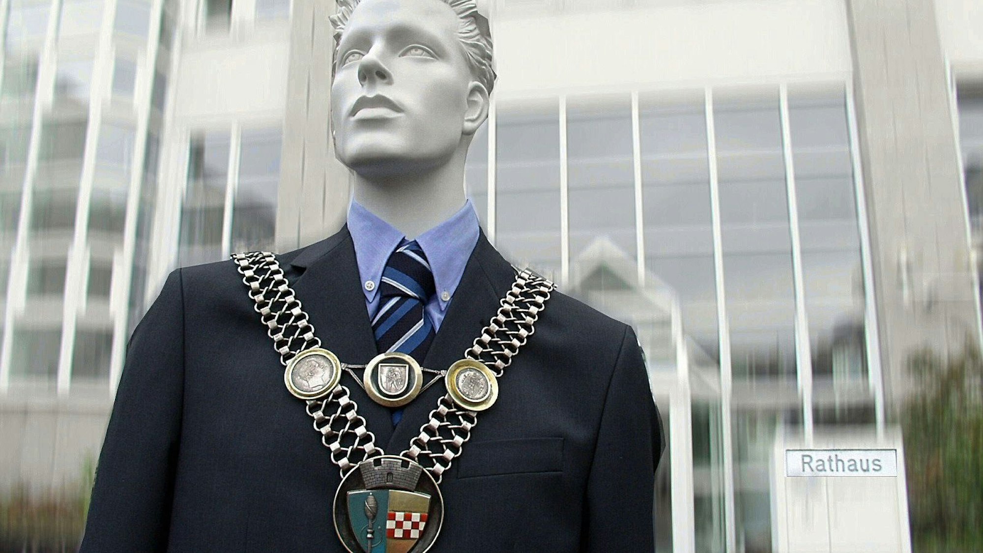 Eine Puppe trägt die Amtskette des Bürgermeisters vor einem Rathaus.