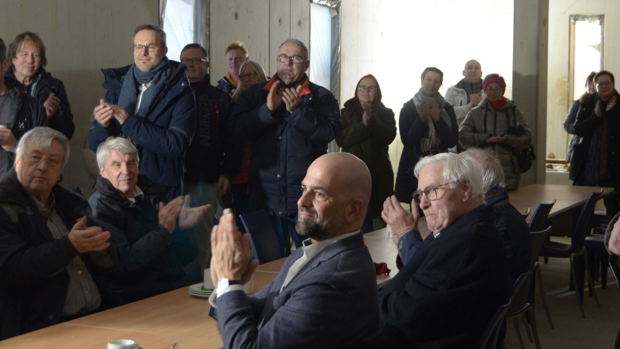 Besucher und Besucherinnen des Richtfestes sitzen und stehen in einem der Räume im Erdgeschoss und klatschen Beifall.