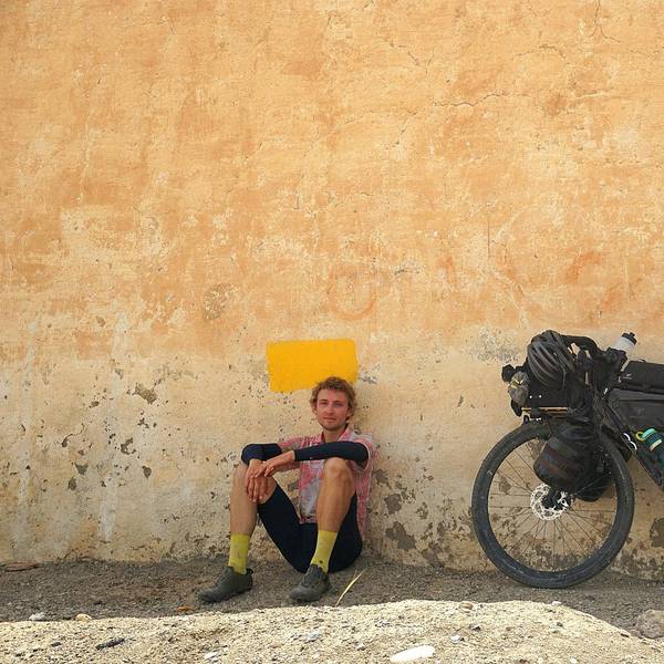 Ein Mann sitzt vor einer Mauer im Schatten, daneben steht sein Fahrrad.