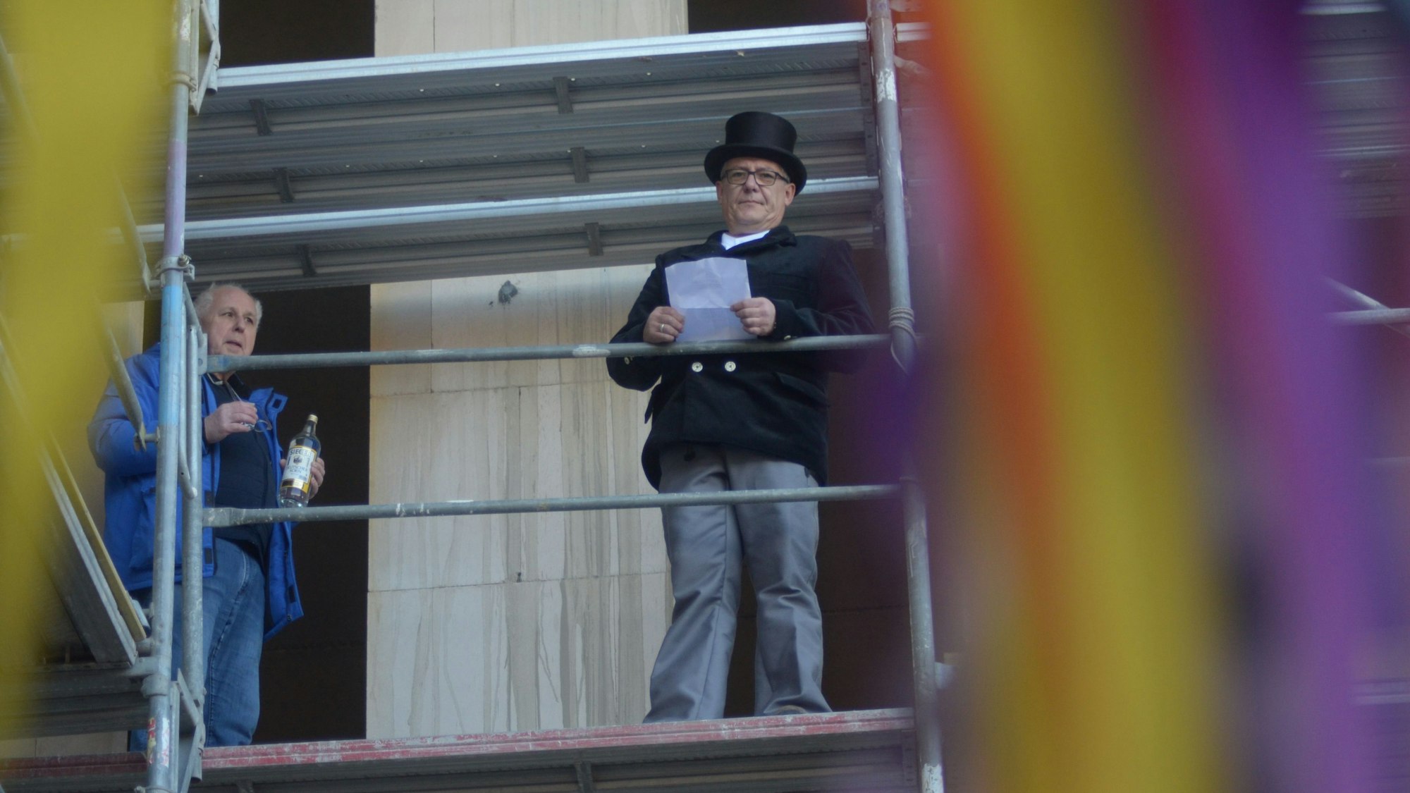 Miguel Coro, mit einem Zylinder auf dem Kopf, und Rolf Kluenter, mit Schnapsflasche, stehen im dritten Obergeschoss auf dem Baugerüst.