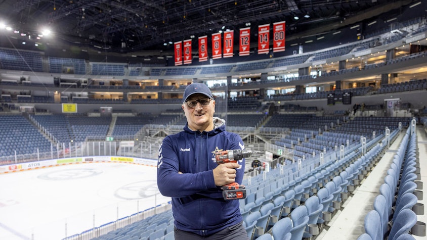 Hausmeister Uzeir Hadzovic steht mit einem Akkuschrauber in der Lanxess-Arena. Im Hintergrund ist noch die Eisfläche zusehen.