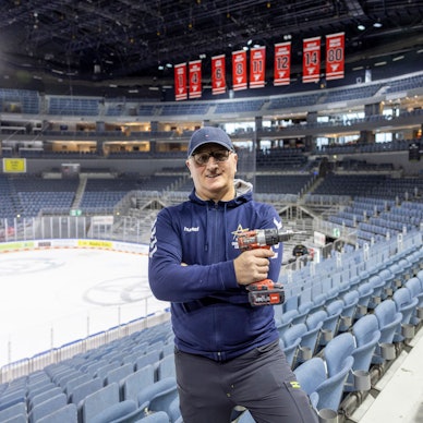 Hausmeister Uzeir Hadzovic steht mit einem Akkuschrauber in der Lanxess-Arena. Im Hintergrund ist noch die Eisfläche zusehen.