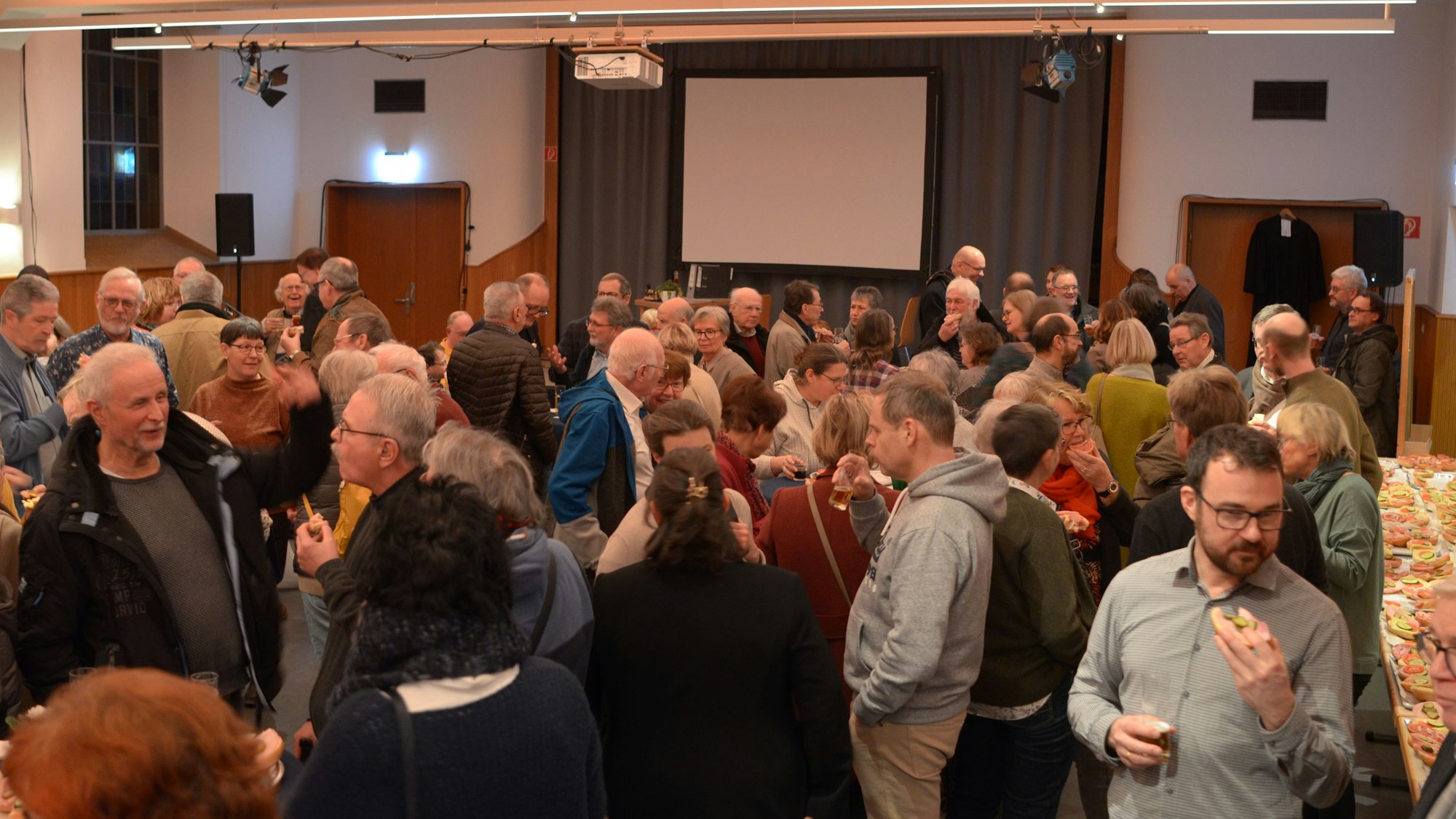 Zahlreiche Gäste stehen im Gemeindesaal der evangelischen Kirche und unterhalten sich, zum Teil mit Gläsern und belegten Brötchen in der Hand.