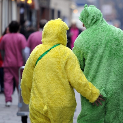 Zwei Menschen in einem gelben und einem grünen Plüsch-Ganzkörperkostüm sind von hinten zu sehen, vor ihnen gehen andere Verkleidete.