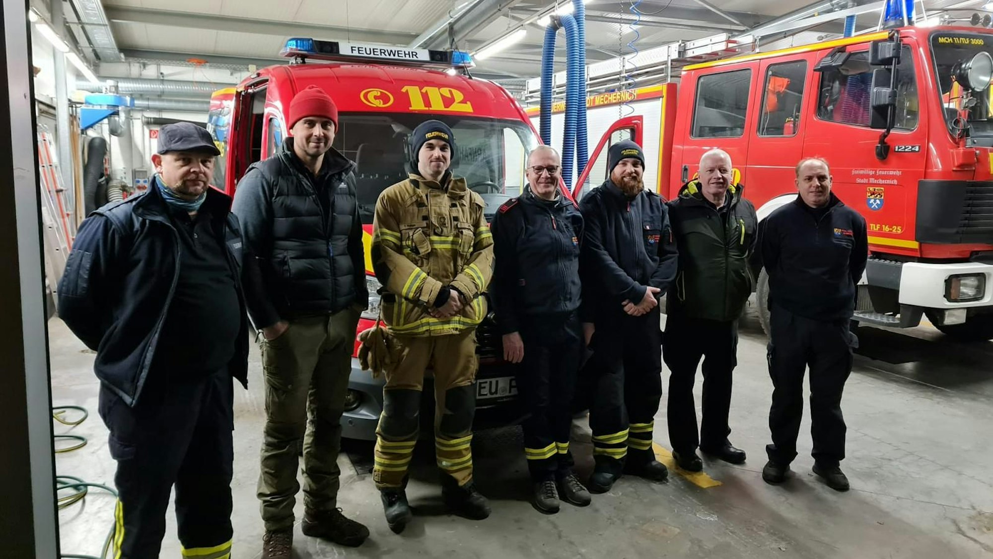 Das Foto zeigt eine Gruppe von Feuerwehrleuten in der Fahrzeughalle der Freiwilligen Feuerwehr Mechernich vor zwei Einsatzfahrzeugen.