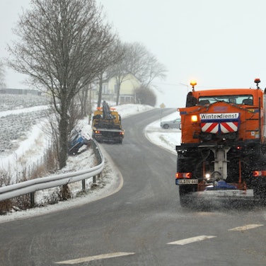 Ein Räumfahrzeug des Winterdienstes befreit eine Straße in der Eifel von Schneemassen. Weitere Schneeflocken fallen vom Himmel. (Symbolbild)