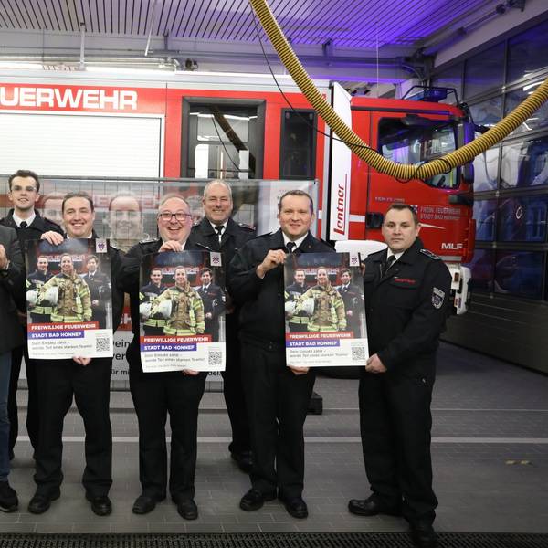 Eine Gruppe von acht Männern steht vor einem Feuerwehrfahrzeug. Sie halten Plakate in die Kamera.