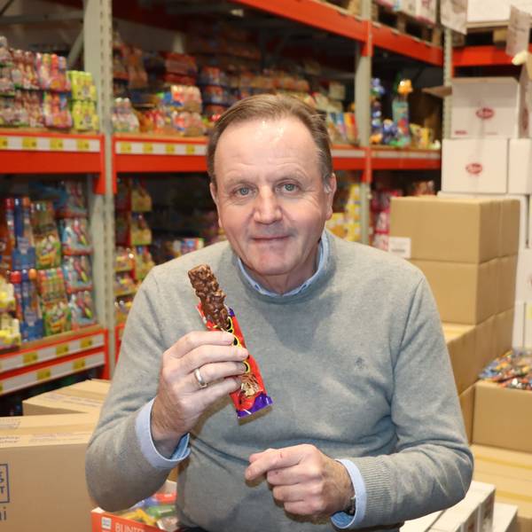 Ein Mann sitzt mit einem Schokoriegel vor Regalen voller Süßigkeiten.