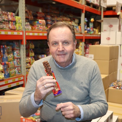 Ein Mann sitzt mit einem Schokoriegel vor Regalen voller Süßigkeiten.