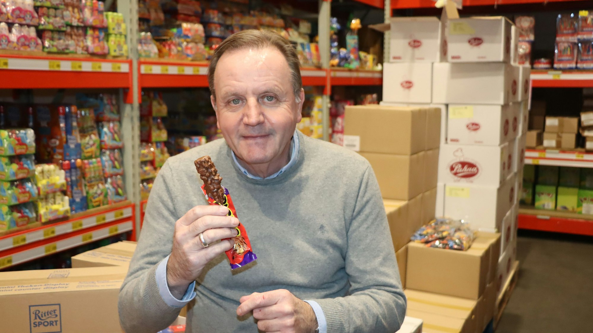 Ein Mann sitzt mit einem Schokoriegel vor Regalen voller Süßigkeiten.
