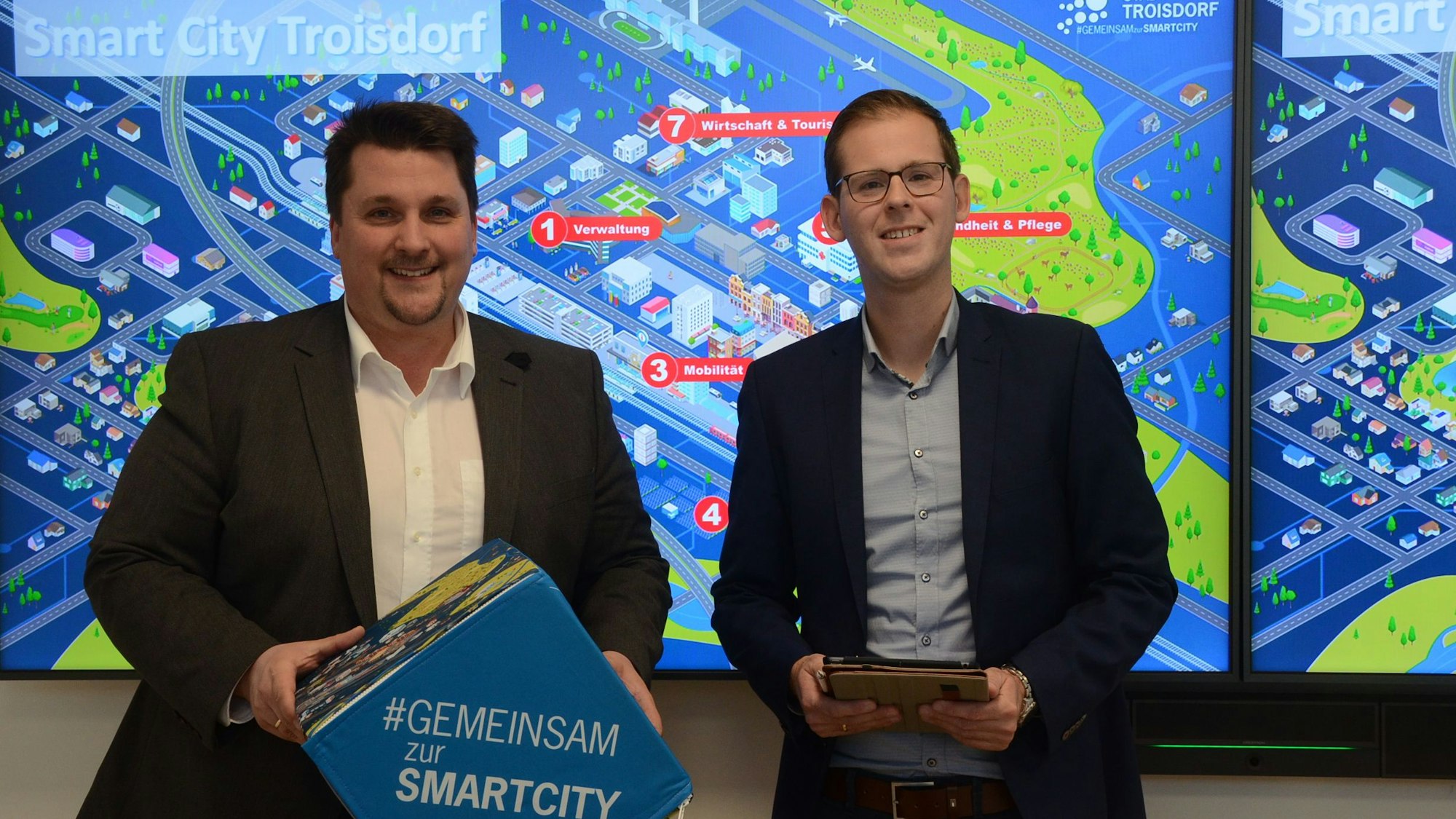 Zwei Männer stehen vor einem bunten Schaubild mit der Überschrift Smart City Troisdorf. Einer der Männer hält einen blauen Würfel, der andere einen Tablet-Computer
