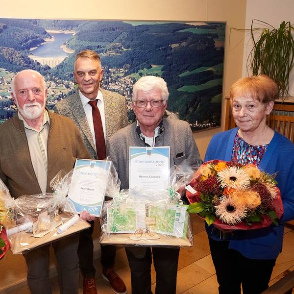 Drei Männer und zwei Frauen stehen vor einem Luftbild von Hellenthal. Die Frauen halten Blumen in den Händen, zwei der Männer Geschenke.
