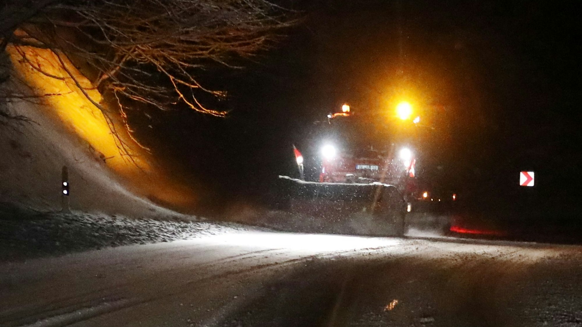 Ein Schneeräumer mit gelbem Rundumlicht fährt durch eine nächtliche Winterlandschaft und räumt Schnee.