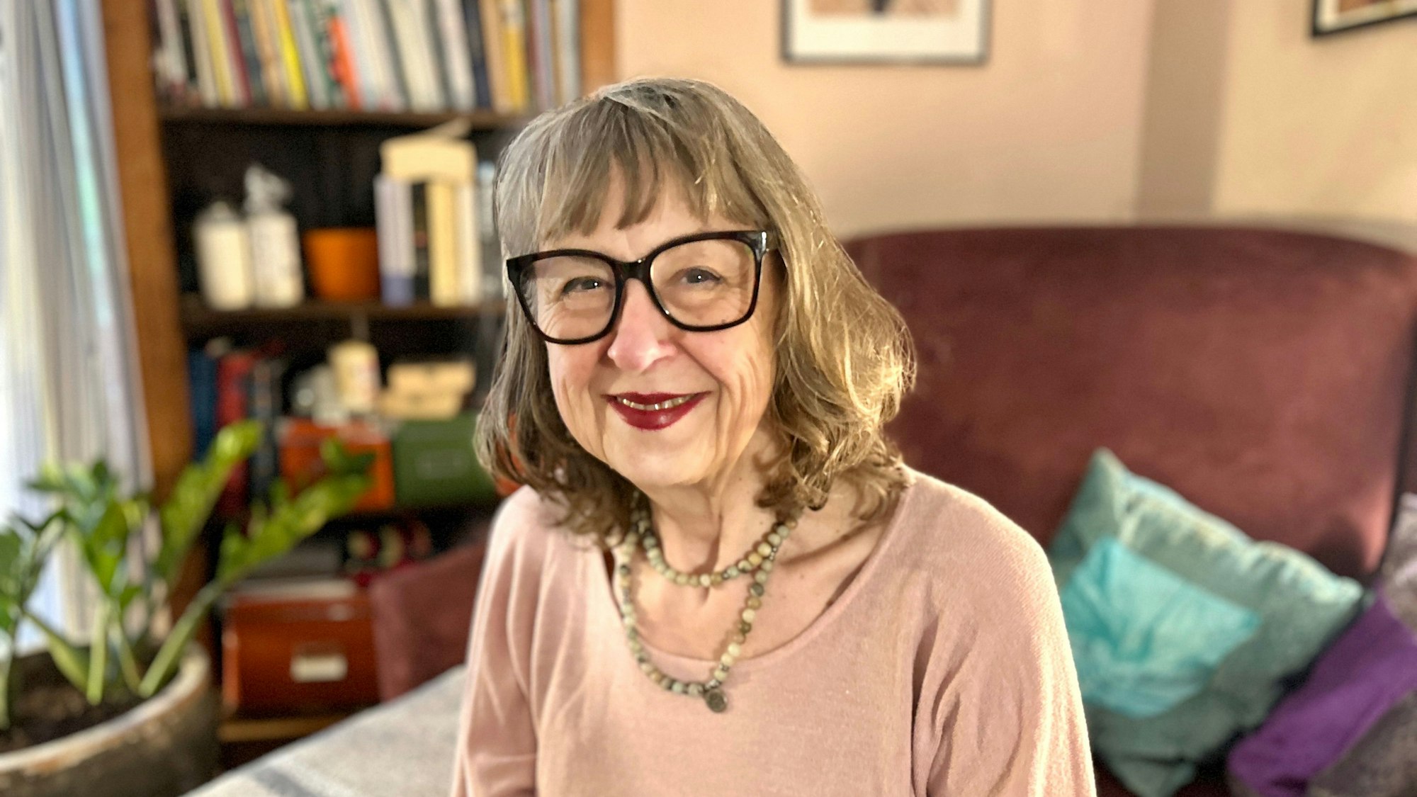 Eine Frau mit schulterlangem Haar und großer Brille sitzt in ihrem Wohnzimmer.