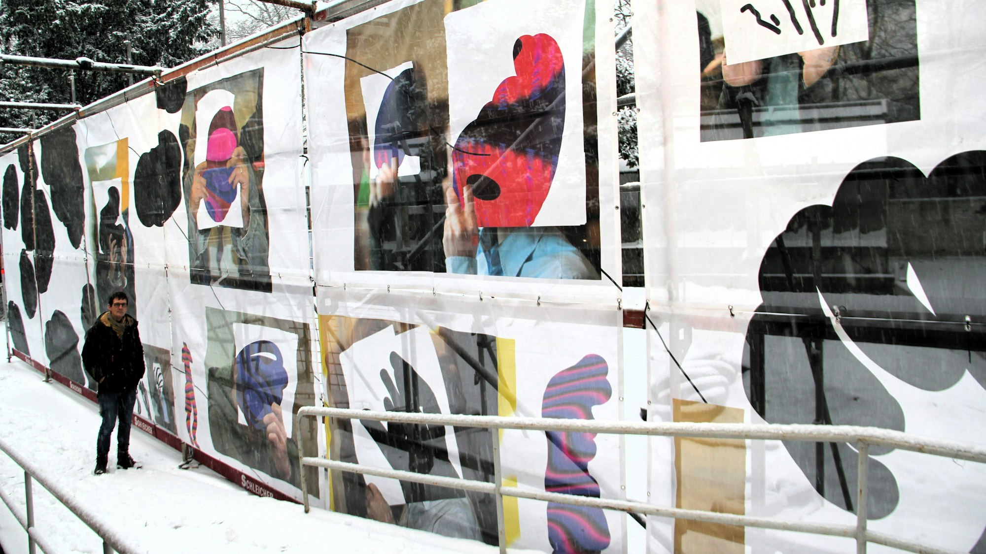 Ein Mann steht an einem riesigen Bauzaun, der mit verschiedenen Bildern dekoriert ist.