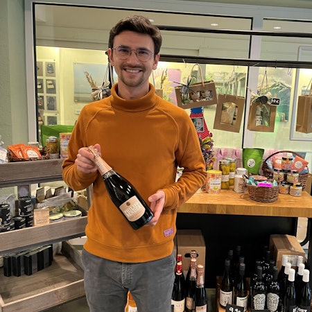 Ein Mann hält eine Flasche Schaumwein in der Hand. Er steht vor Regalen mit hochwertigen Lebensmitteln.