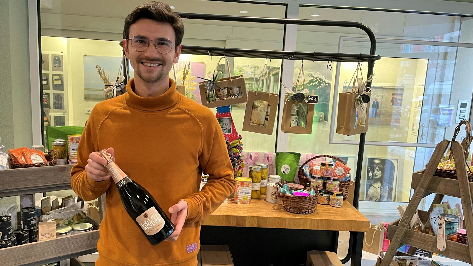 Ein Mann hält eine Flasche Schaumwein in der Hand. Er steht vor Regalen mit hochwertigen Lebensmitteln.