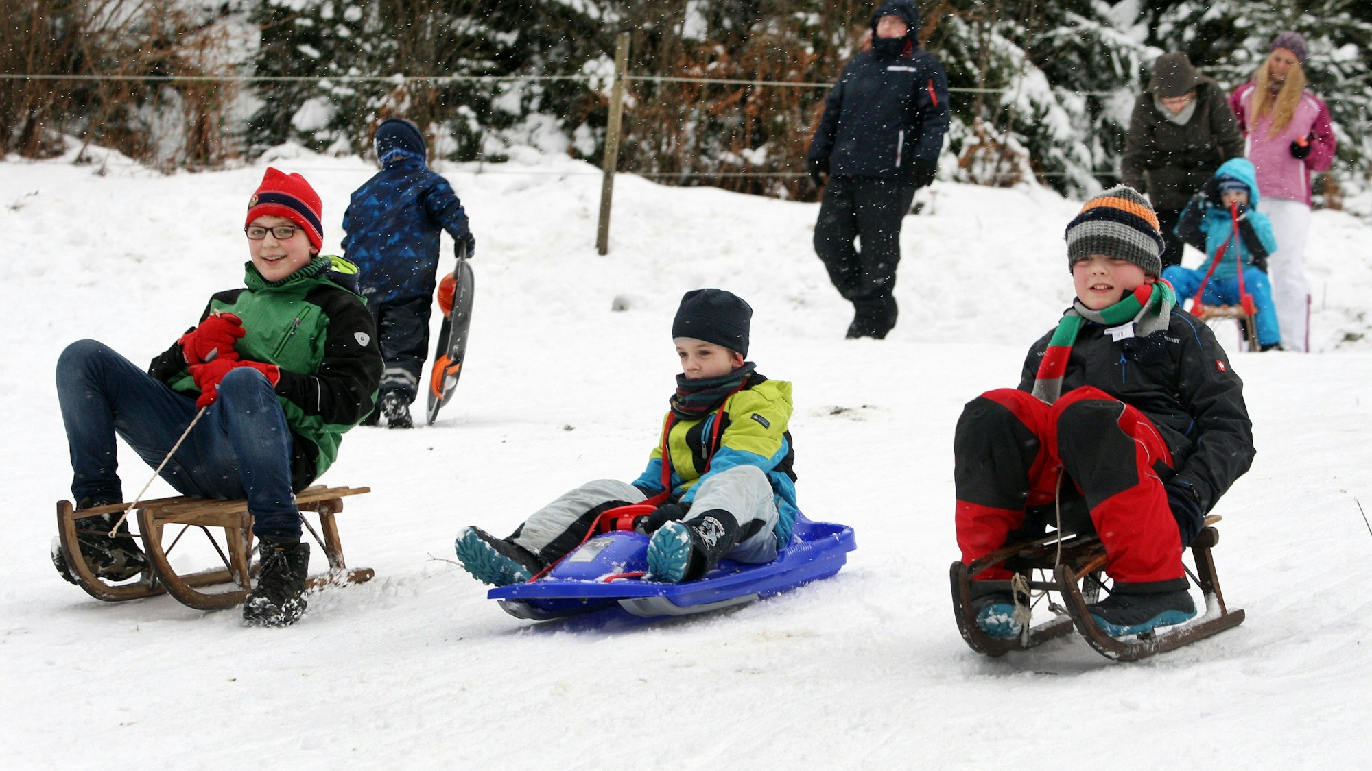 Drei Kinder fahren auf Schlitten und einem Bob einen Abhang hinunter.