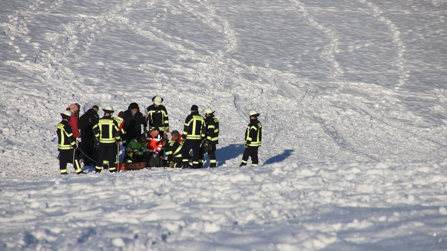 Rettungskräfte stehen auf einem tief verschneiten Hang. In ihrer Mitte knien Helfer und versorgen eine Patientin.