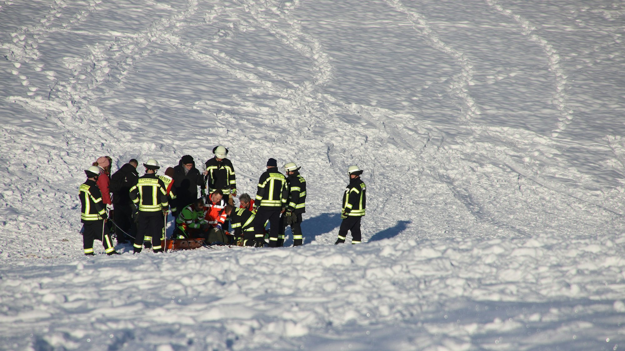 Rettungskräfte stehen auf einem tief verschneiten Hang. In ihrer Mitte knien Helfer und versorgen eine Patientin.