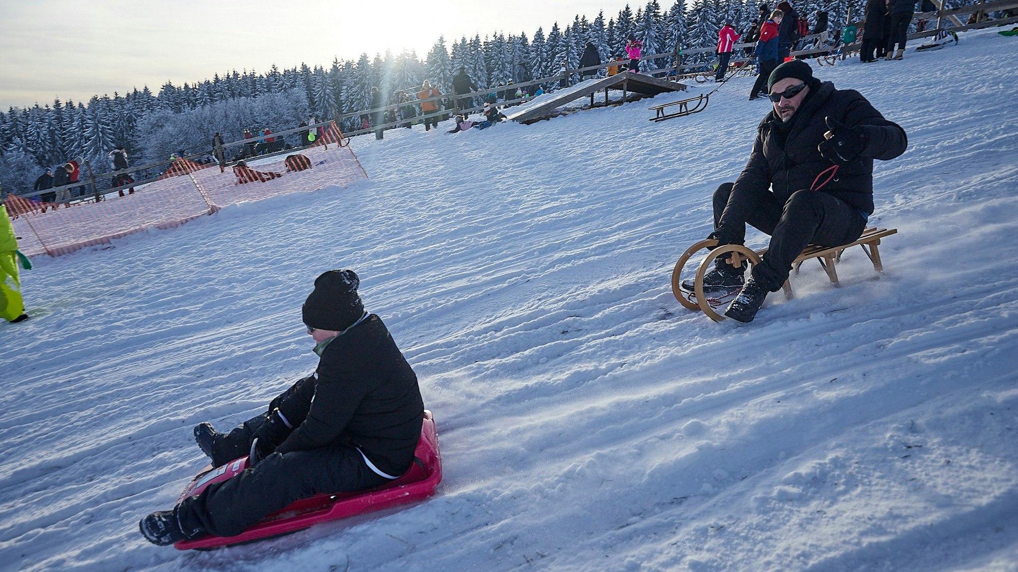 Ein Junge saust auf einem Plastikbob den Hang am Weißen Stein hinunter, dahinter ein Mann auf einem Schlitten.