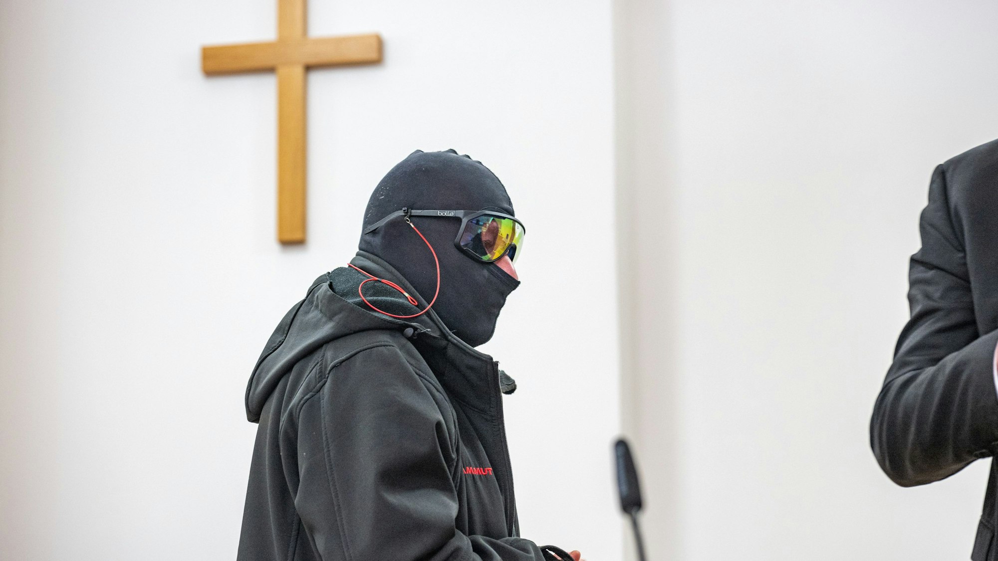 Ein Man in schwarzem Anorak, mit Sturmhaube und verspiegelter Sonnenbrille steht in einem Raum, an der Wand hängt ein Holzkreuz.