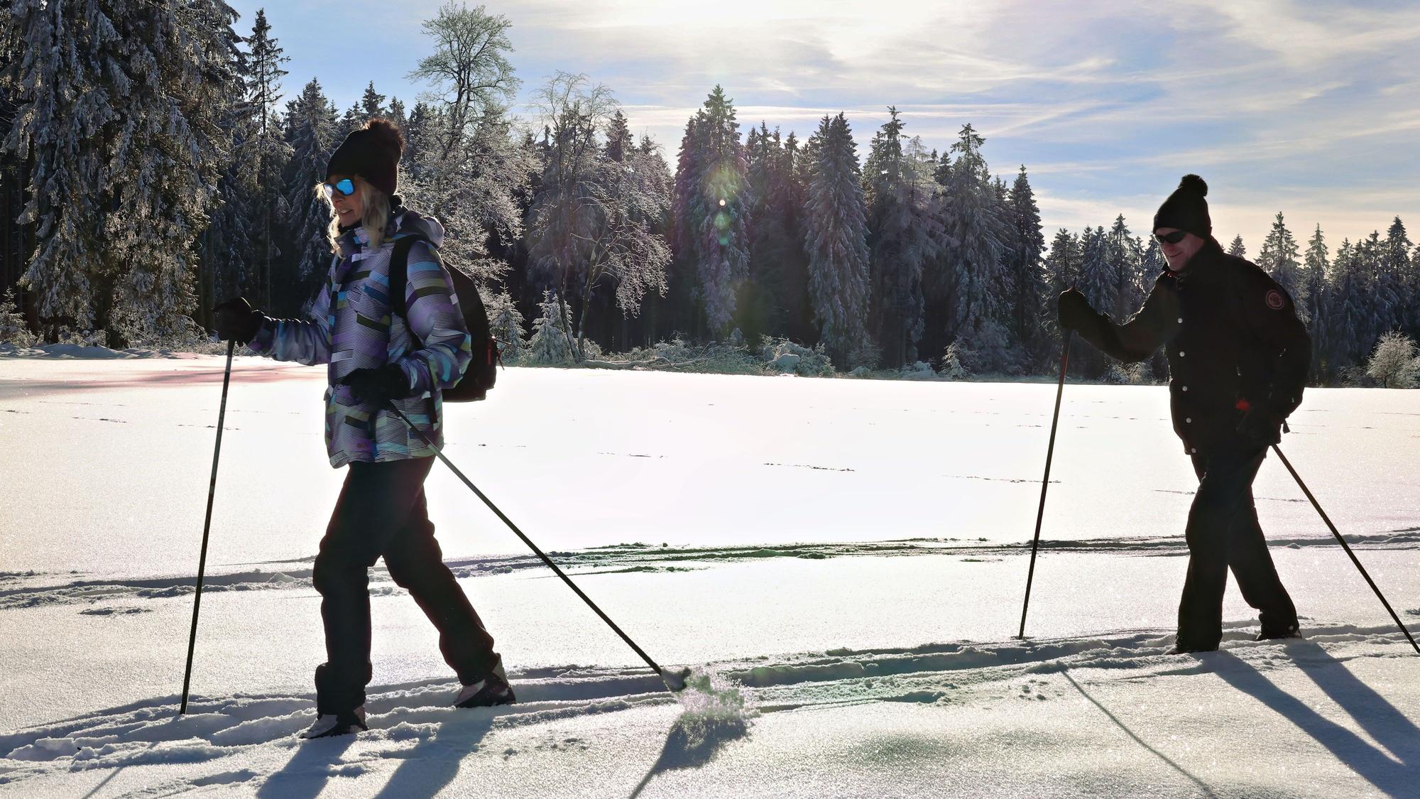 Ein junges Pärchen ist auf Skiern im glitzernden Pulverschnee unterwegs, im Hintergrund verschneite Wälder.