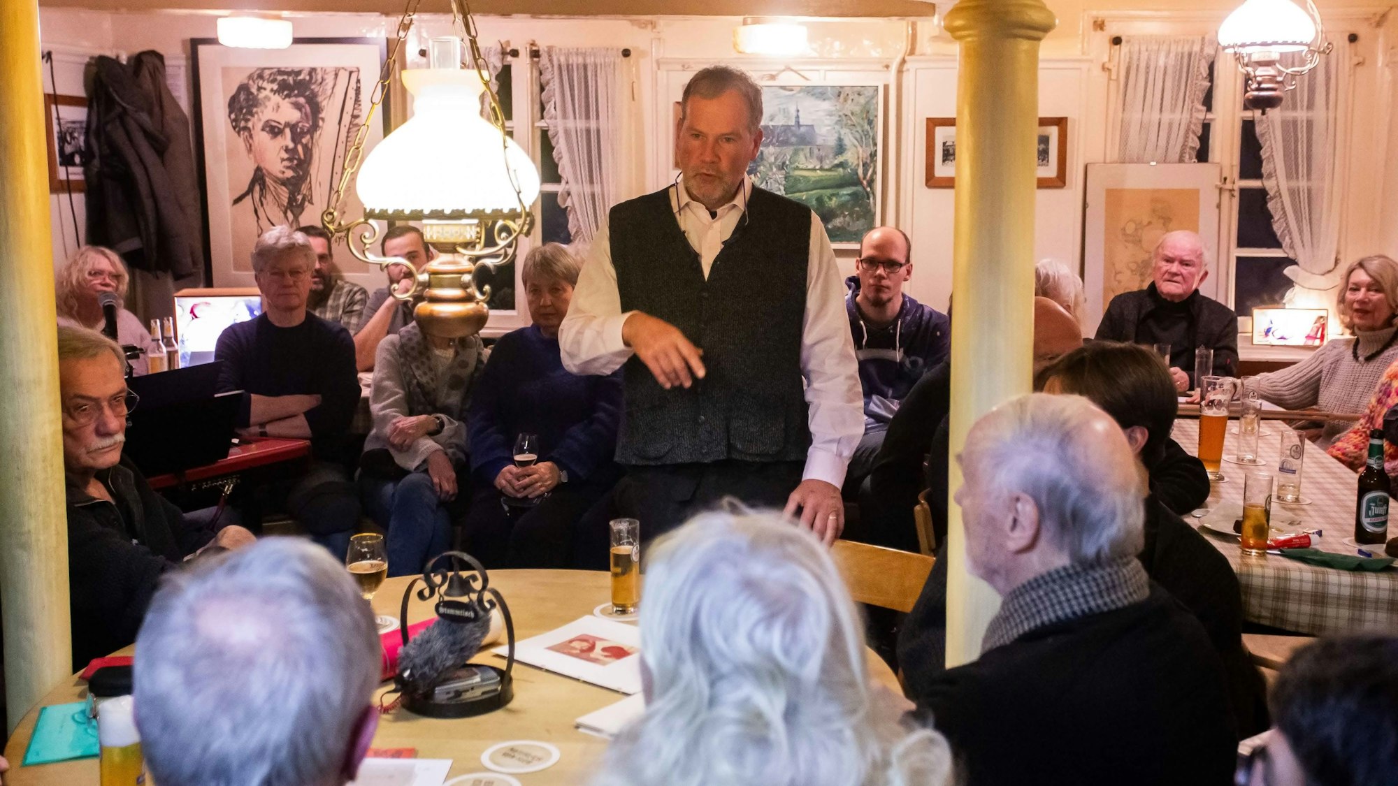 Ein Mann steht in einer dicht besetzten Kneipe und spricht zu den Umsitzenden.