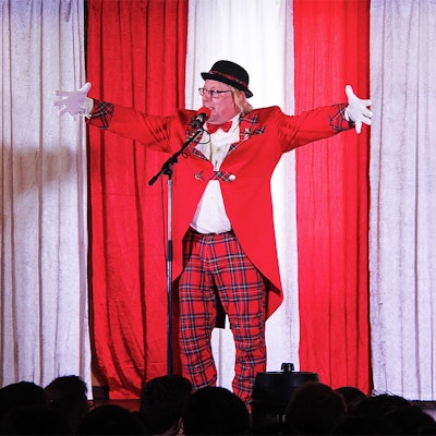 Der Redner Stefan van der Eerdwich steht gestikulierend auf der rot-weiß dekorierten Bühne. Er trägt einen schwarzen Hut, einen roten Frack und eine rot-schwarz karierte Hose.