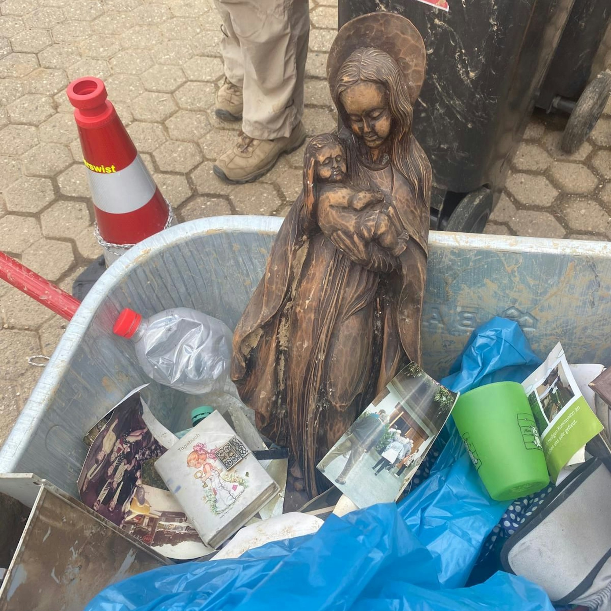 Eine holzgeschnitzte Madonnenfigur liegt in einer Schubkarre zwischen Plastikflaschen, Fotografien und einem Tagebuch.