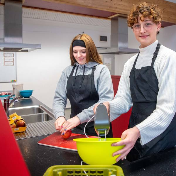 Das Bild zeigt Celina und Jakob in der Schulküche der Zülpicher Hauptschule. Celina schneidet einen Apfel, Jakob hat einen Mixer in der Hand.