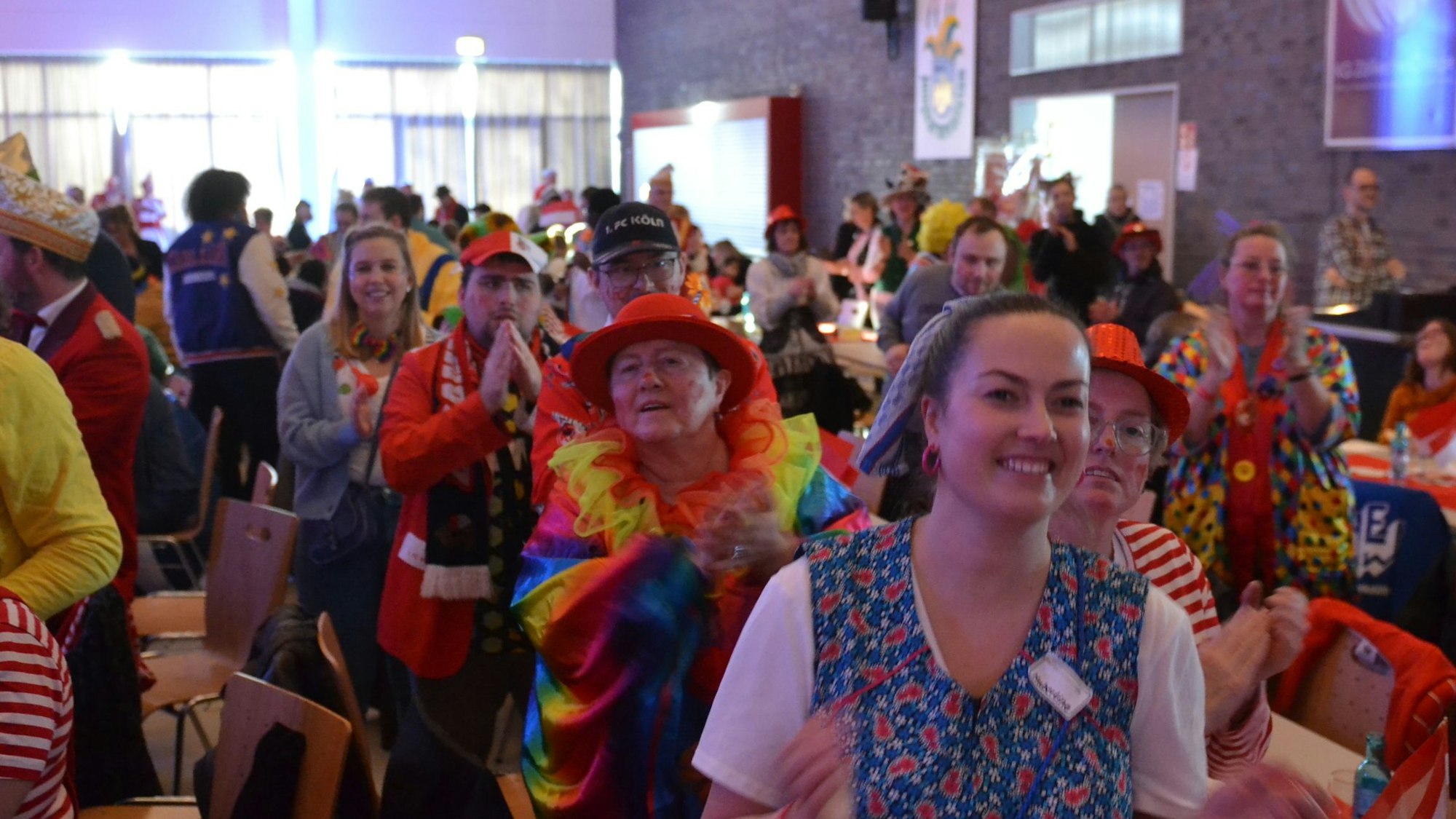 Kostümierte Jecke haben sich von ihren Stühlen erhoben, sie klatschen und lachen.