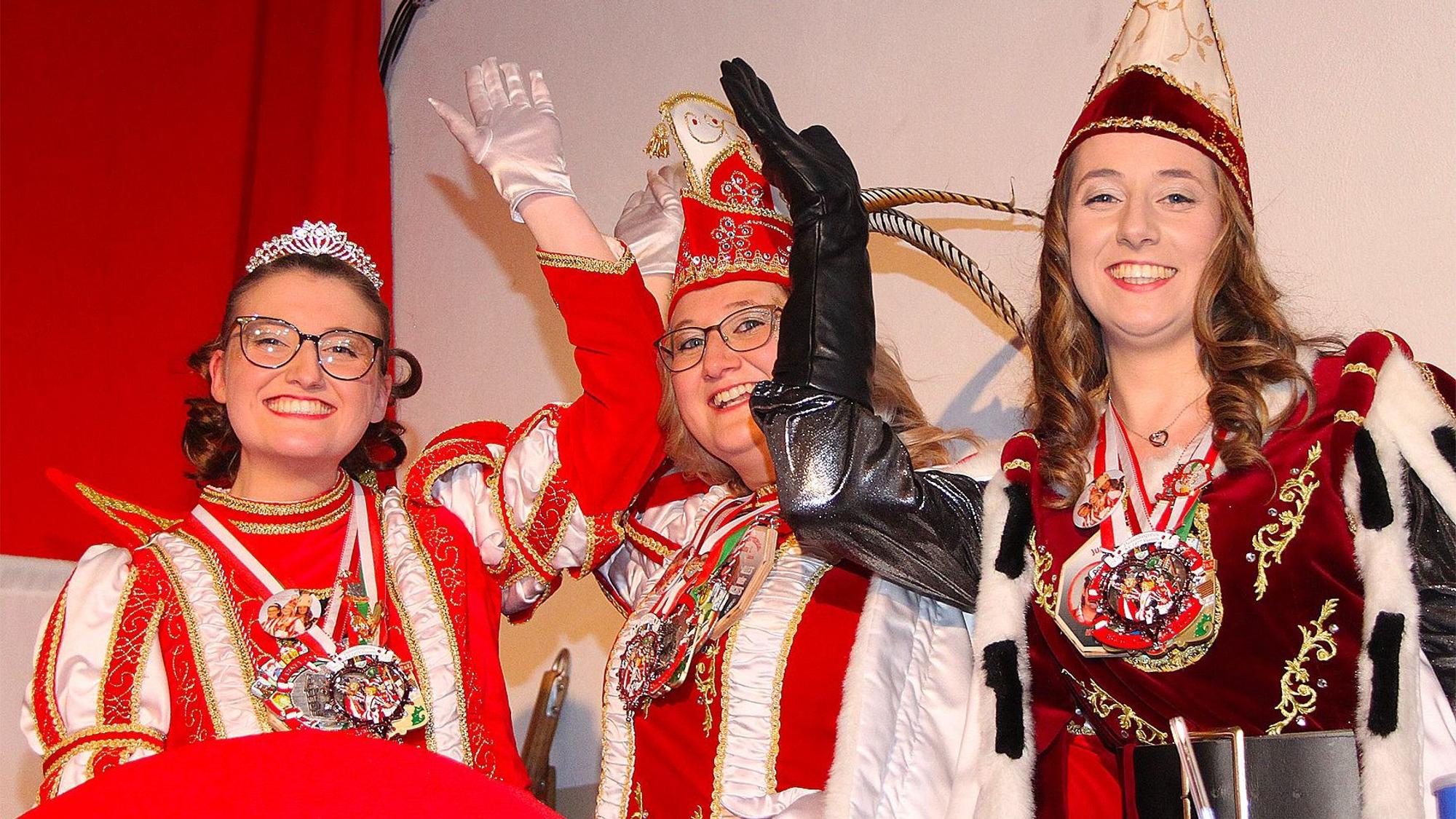Die drei Frauen, die das Dreigestirn bilden, lachen fröhlich in die Kamera. Sie tragen jeweils mehrere Karnevalsorden.