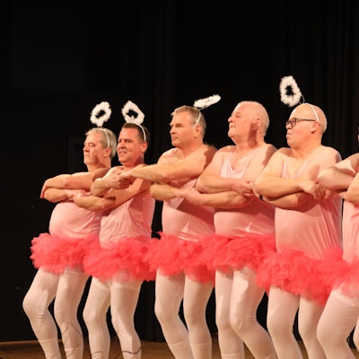 Mehrere Männer tragen rosa Tütüs und tanzen in einer Reihe. Auf dem Kopf tragen sie Heiligenscheine.