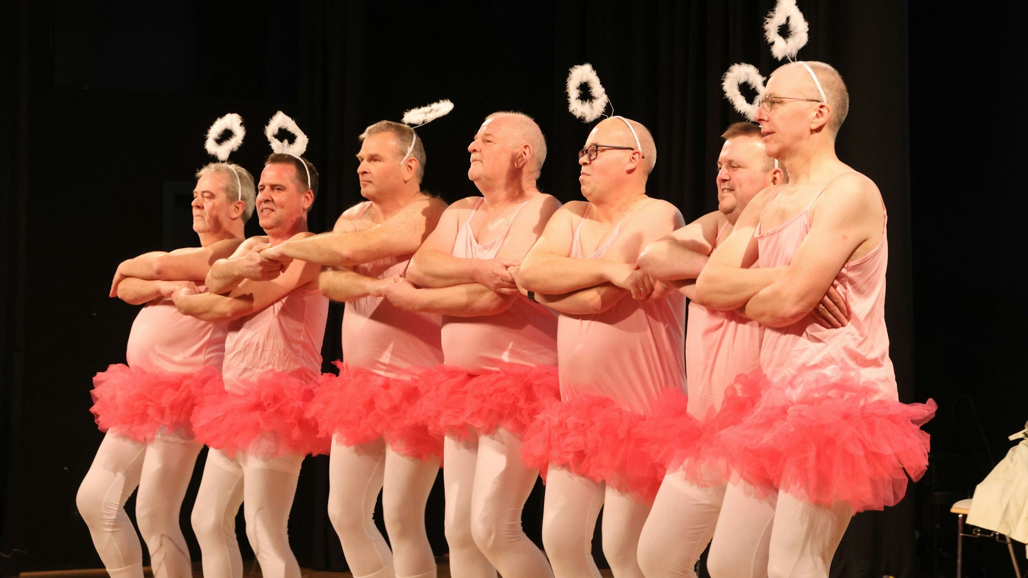 Mehrere Männer tragen rosa Tütüs und tanzen in einer Reihe. Auf dem Kopf tragen sie Heiligenscheine.