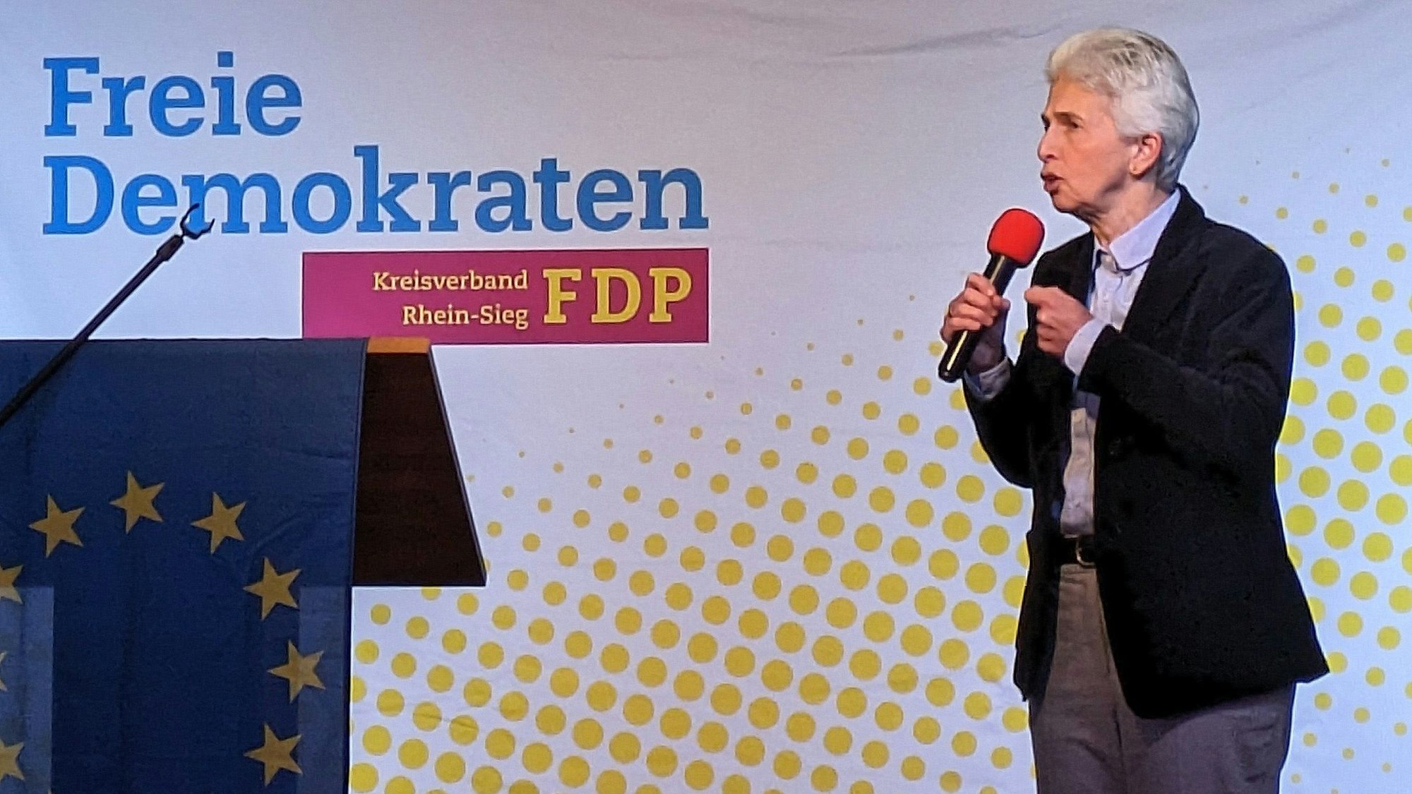Eine Frau mit kurzen grauen Haaren, schwarzem Sakko und grauer Hose spricht in ein Mikrofon. Auf der Wand steht Freie Demokraten.